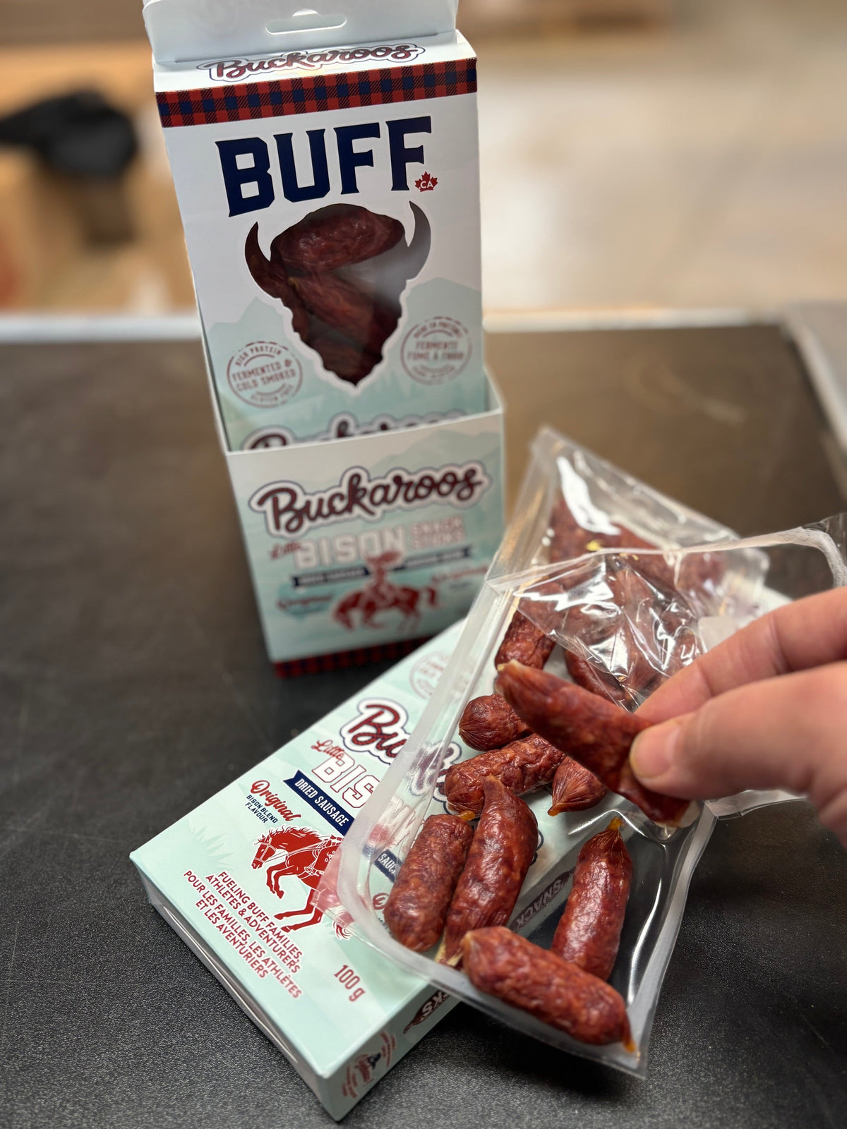 Hand holding BUFF Buckaroos mini bison sausage snack sticks from an open 100 g box on a counter