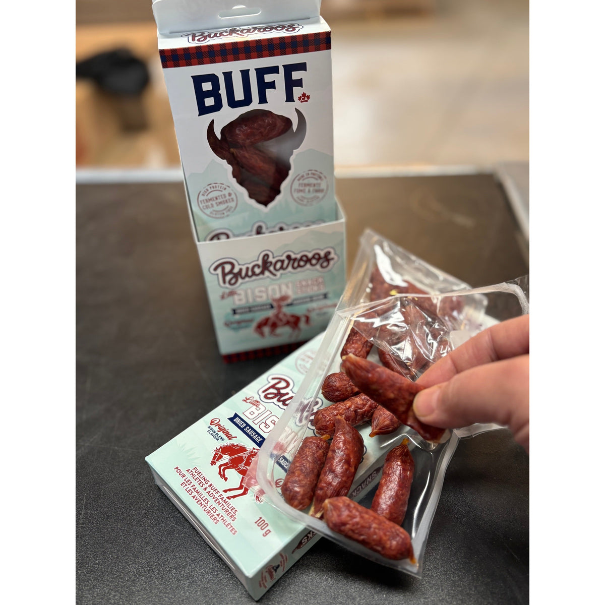 Hand holding BUFF Buckaroos mini bison sausage snack sticks from an open 100 g box on a counter